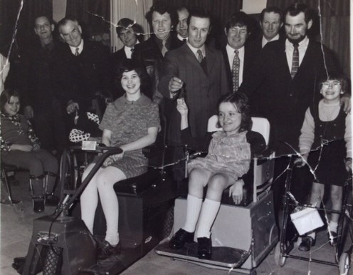 Black and white photograph of 3 disabled girls smiling at the camera. A group of ten suited men standing behind them, looking quite paternalistic.